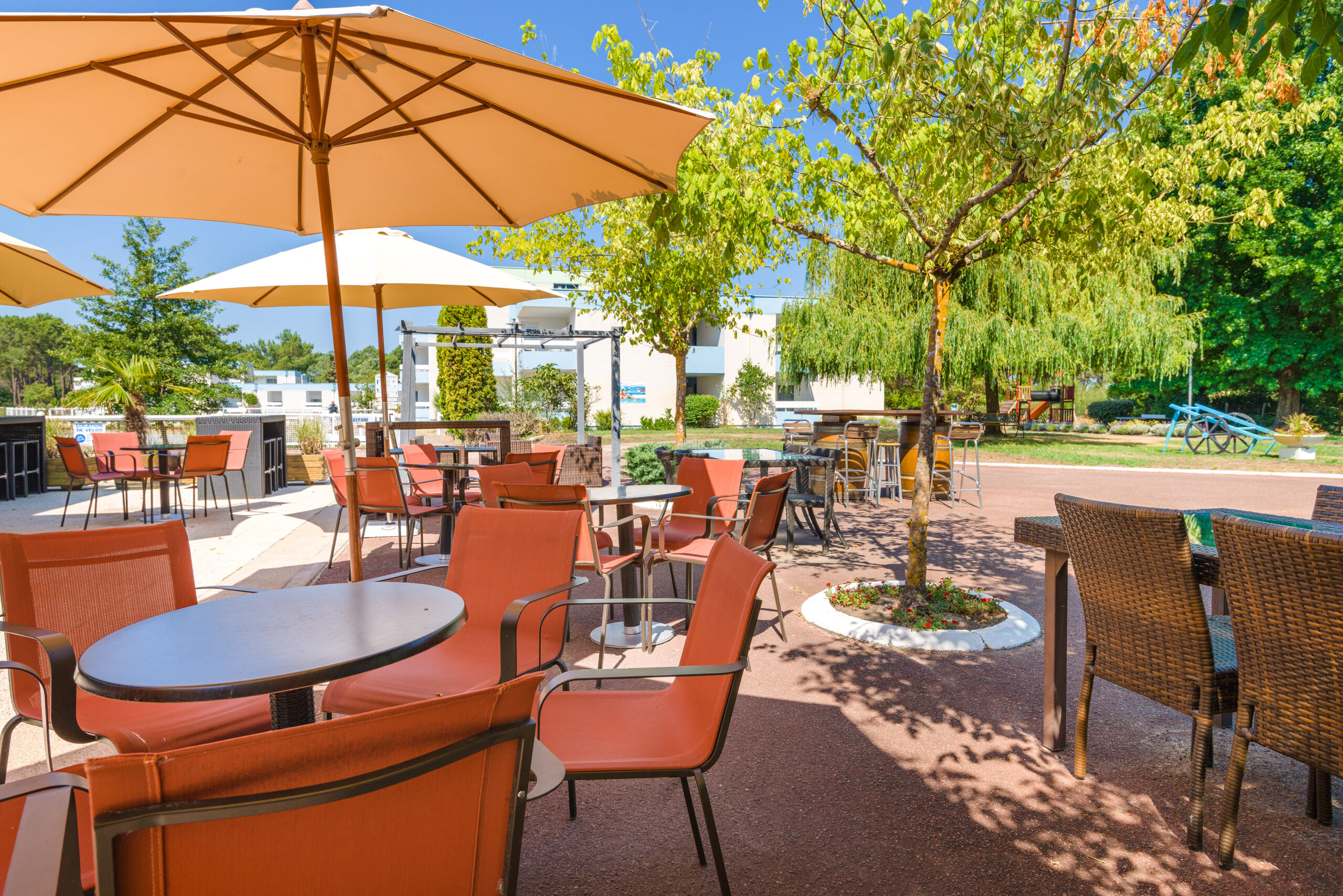 Terrasse extérieure ombragée avec tables et chaises, vue sur un jardin arboré dans un village de vacances en France.