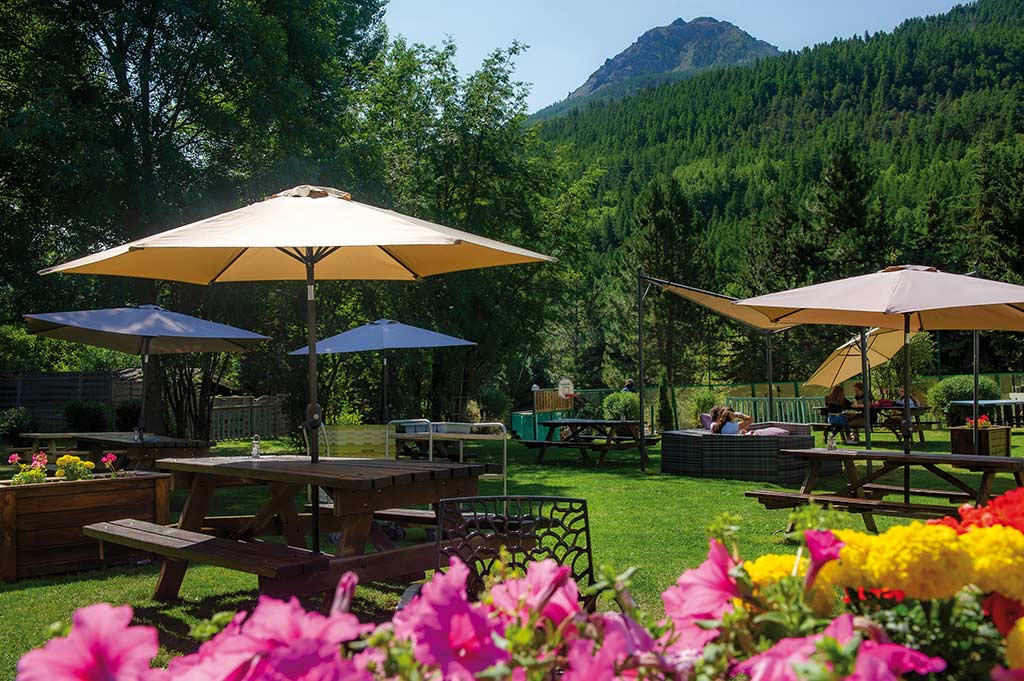 Terrasse extérieure fleurie d'un Village Vacances Azureva en montagne avec tables de pique-nique, parasols et vue panoramique sur les sommets boisés