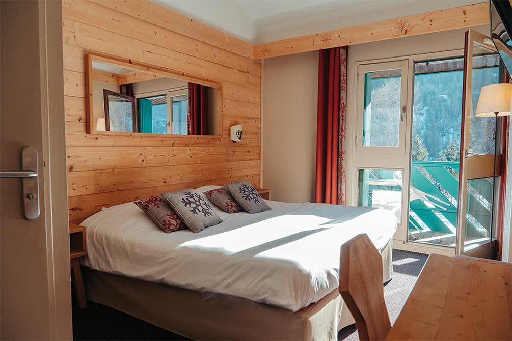 Chambre confortable en montagne avec décoration bois chaleureux, grand lit et balcon avec vue sur la forêt enneigée