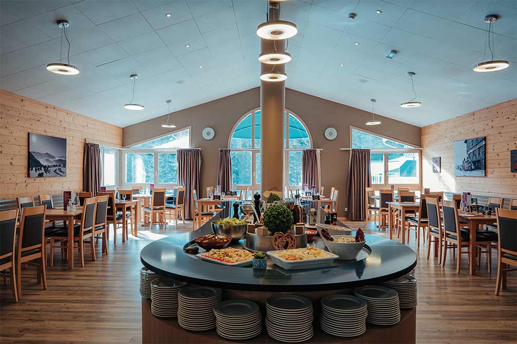 Salle de restaurant spacieuse en montagne avec buffet central, grandes baies vitrées cintrées et décoration bois chaleureux