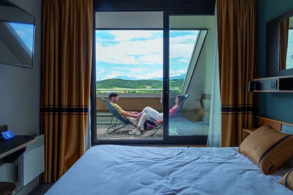 Chambre Azureva avec vue sur terrasse privée où deux personnes se détendent sur des transats face à un panorama de montagnes verdoyantes