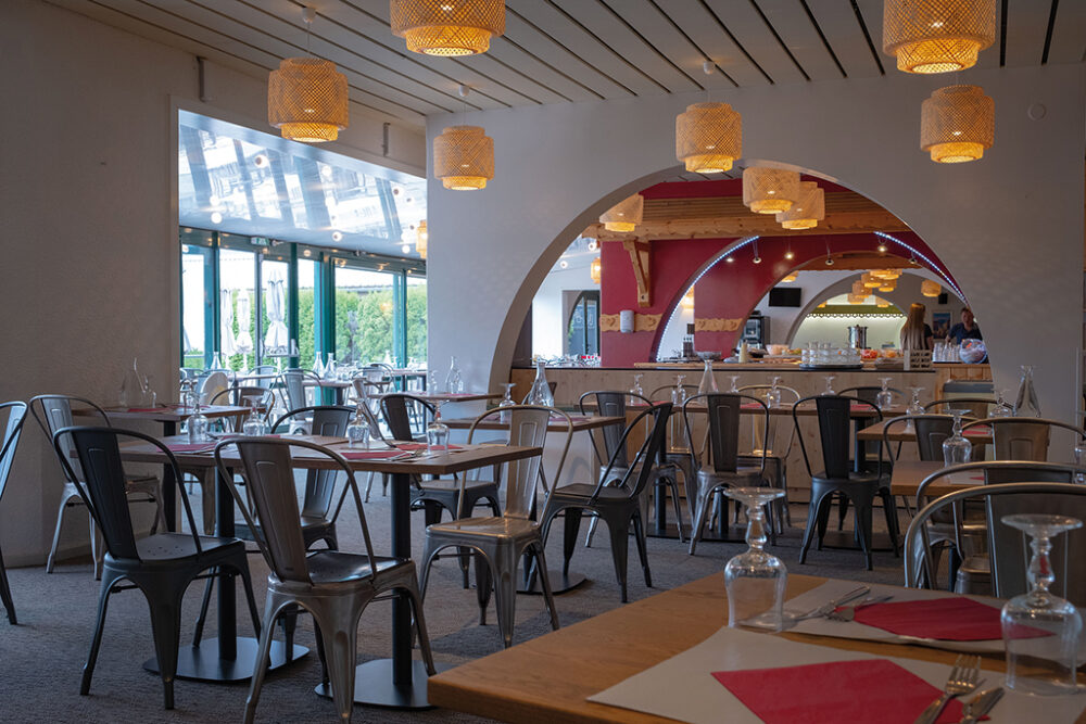 Salle de restaurant vacances bleues spacieuse et lumineuse avec décoration moderne, luminaires en rotin et arches donnant sur la cuisine ouverte