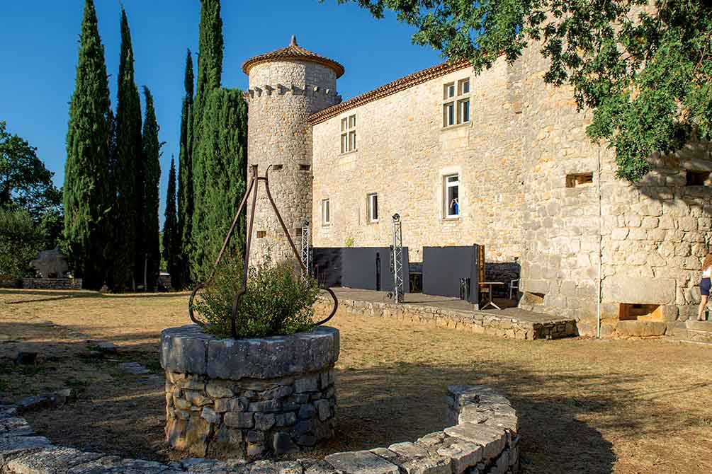 Château en pierre avec tour et architecture médiévale abritant un établissement, entouré de cyprès et d'un puits ancien sous le soleil du Sud