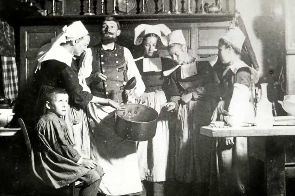Photographie ancienne montrant une scène de vie traditionnelle en Bretagne avec des femmes en coiffe et un homme en costume préparant un repas