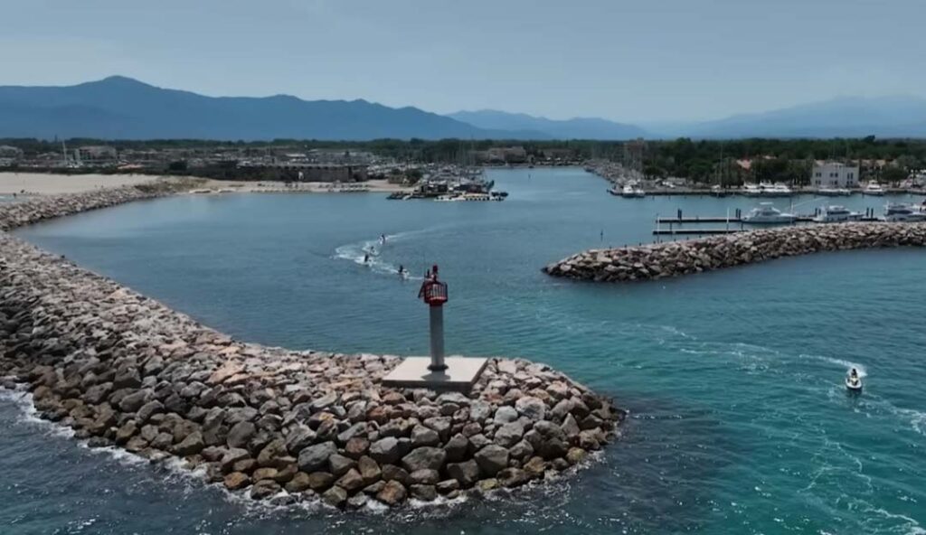 Entrée de port méditerranéen avec petit phare rouge sur jetée, eaux turquoise et montagnes en arrière-plan dans les Pyrénées-Orientales