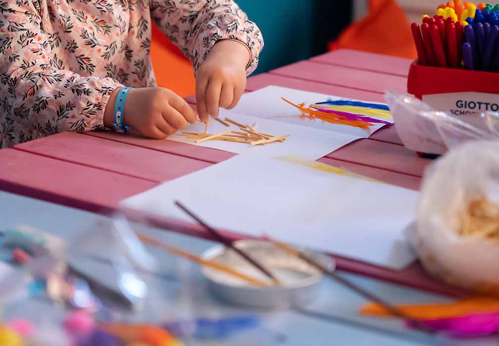 Gros plan sur les mains d'un enfant faisant une activité manuelle avec des brindilles et des plumes, dans un club enfant.