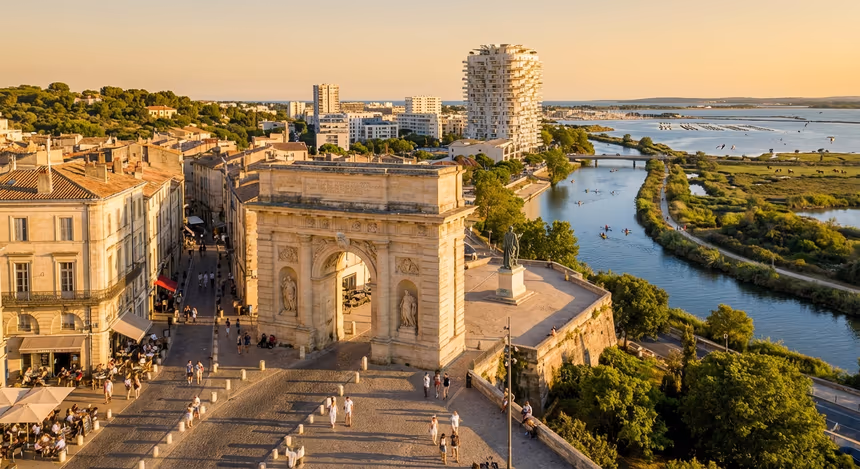 Vue ultra réaliste du centre historique de Montpellier au coucher de soleil, avec l’arc de triomphe, l’esplanade du Peyrou, les ruelles de l’Écusson, le Lez avec des kayaks, la garrigue, des zones protégées comme l’étang de l’Or, et en arrière-plan des quartiers modernes comme Port Marianne et l’Arbre Blanc à La Grande-Motte