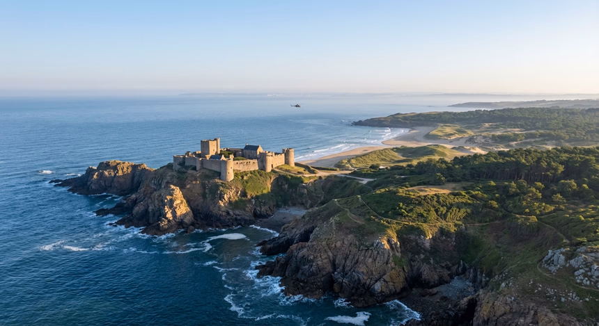 Vue matinale ultra-réaliste de la côte de l’Île d’Yeu avec un hélicoptère survolant la mer, dunes sableuses, falaises rocheuses et le Vieux Château du XIVe siècle sur un éperon rocheux